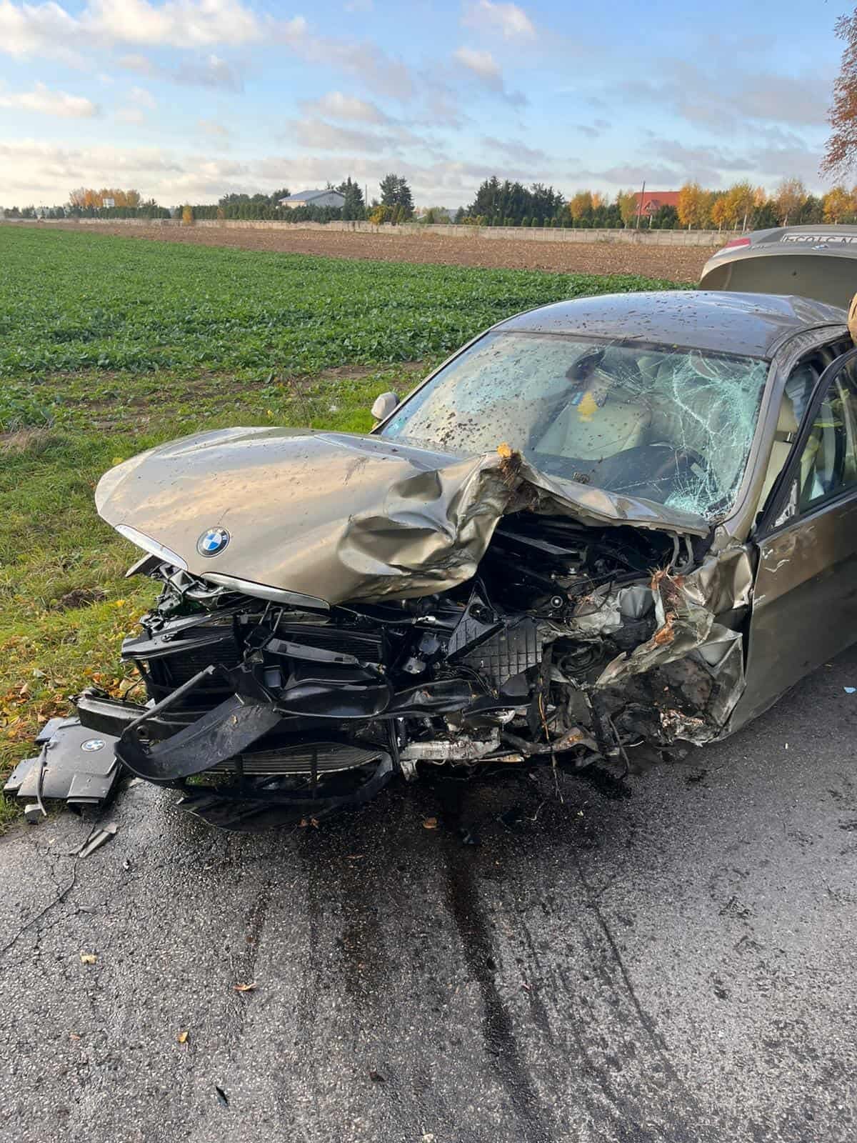 Uszkodzony samochód osobowy leży na poboczu drogi, z widocznymi uszkodzeniami przodu i rozbitym przednią szybą, w tle pola.