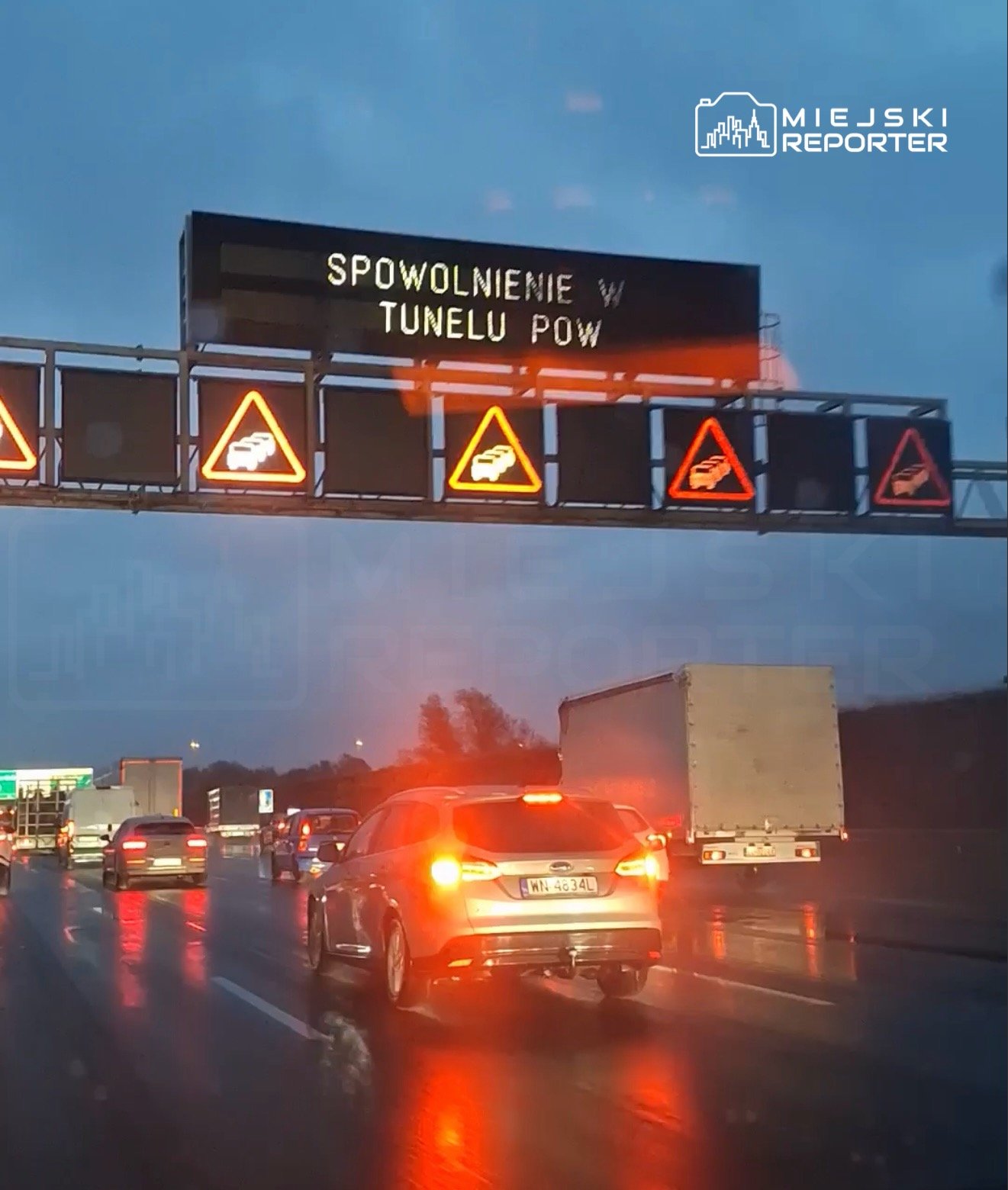 Na drogowskazie wyświetla się komunikat "SPÓWOLNIENIE W TUNELU POW", a w tle widać ruch pojazdów na deszczowej jezdni.