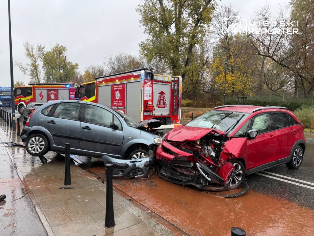 Dwa uszkodzone samochody osobowe po kolizji na ulicy, obok stoją wozy strażackie. Deszczowa pogoda, kałuże na jezdni.