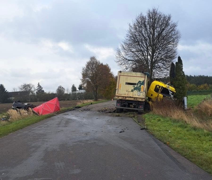 Złomowany żółty ciężarówka w rowie, obok przewrócony czerwony namiot na wiejskiej drodze otoczonej drzewami.