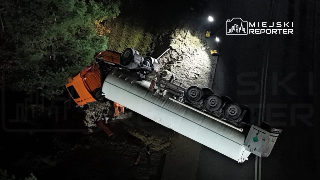 Ciągnik siodłowy przewrócony na poboczu drogi, otoczony przez drzewa, w nocy oświetlony przez latarnie.