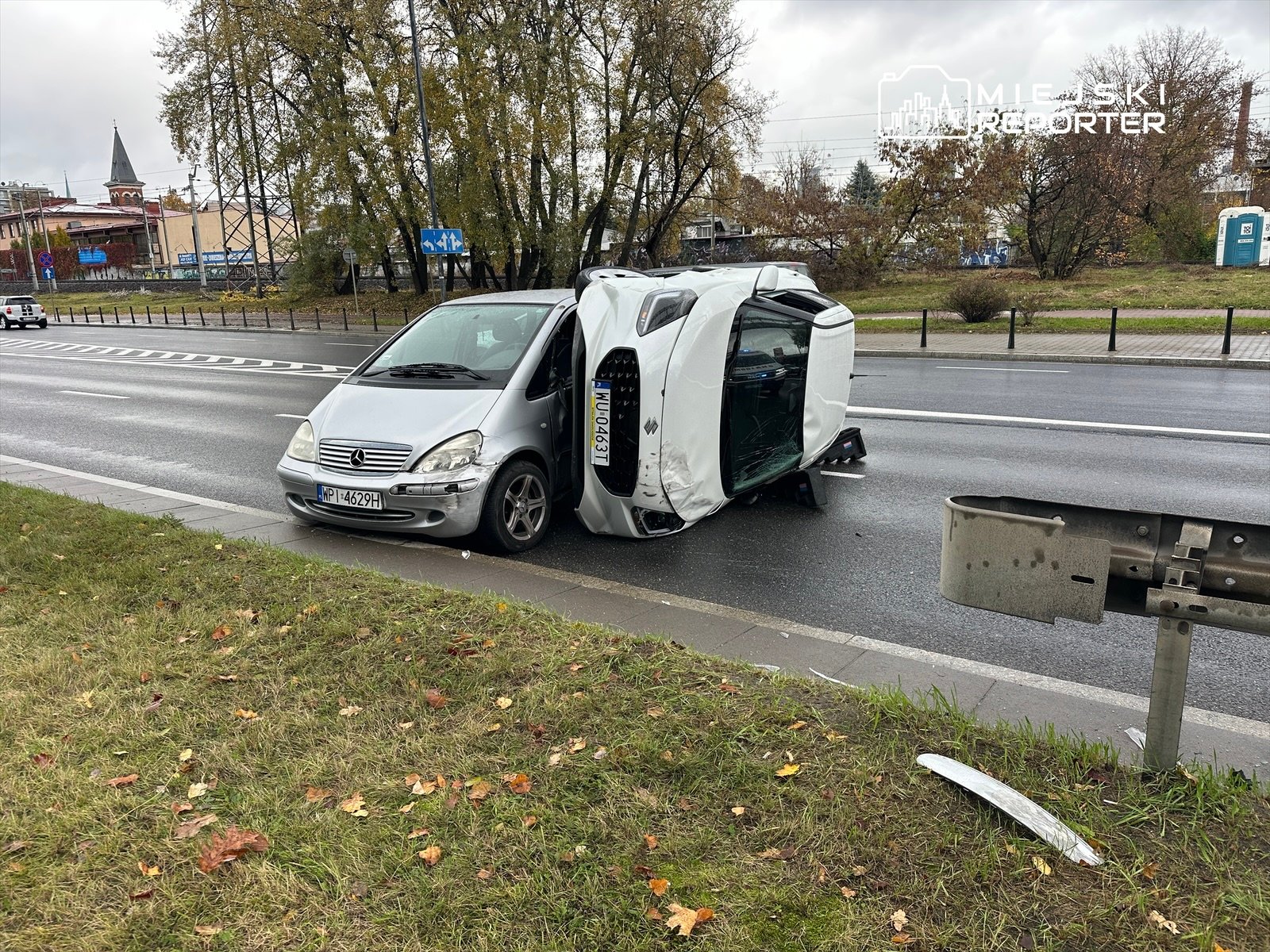 Samochód osobowy leży na boku na poboczu drogi, obok przejeżdżające inne pojazdy. W tle widać budynki i drzewa.