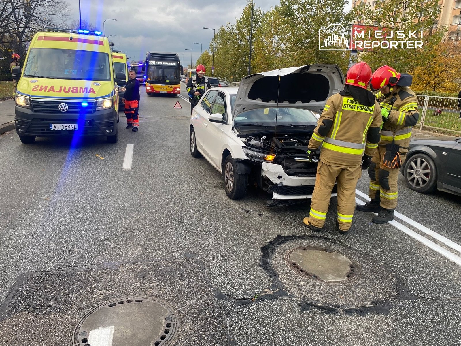 Dwie karetki pogotowia oraz strażacy sprawdzają uszkodzony samochód osobowy na ruchliwej ulicy.