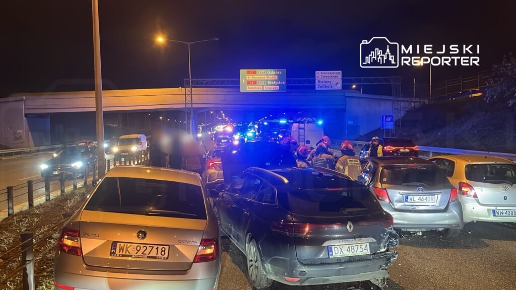 Grupa ratowników w hełmach interweniuje na drodze, obok zderzonych samochodów w nocy. W tle widać policyjne światła.