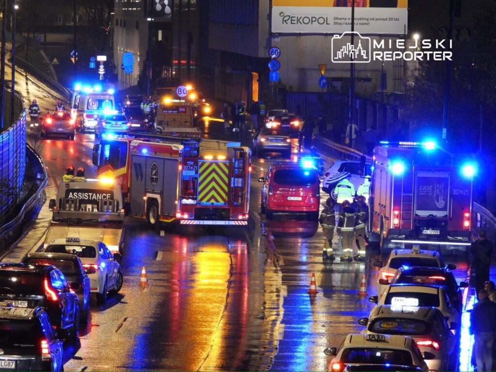 Na mokrej jezdni, w otoczeniu pojazdów ratunkowych, strażacy i ratownicy medyczni udzielają pomocy poszkodowanym.