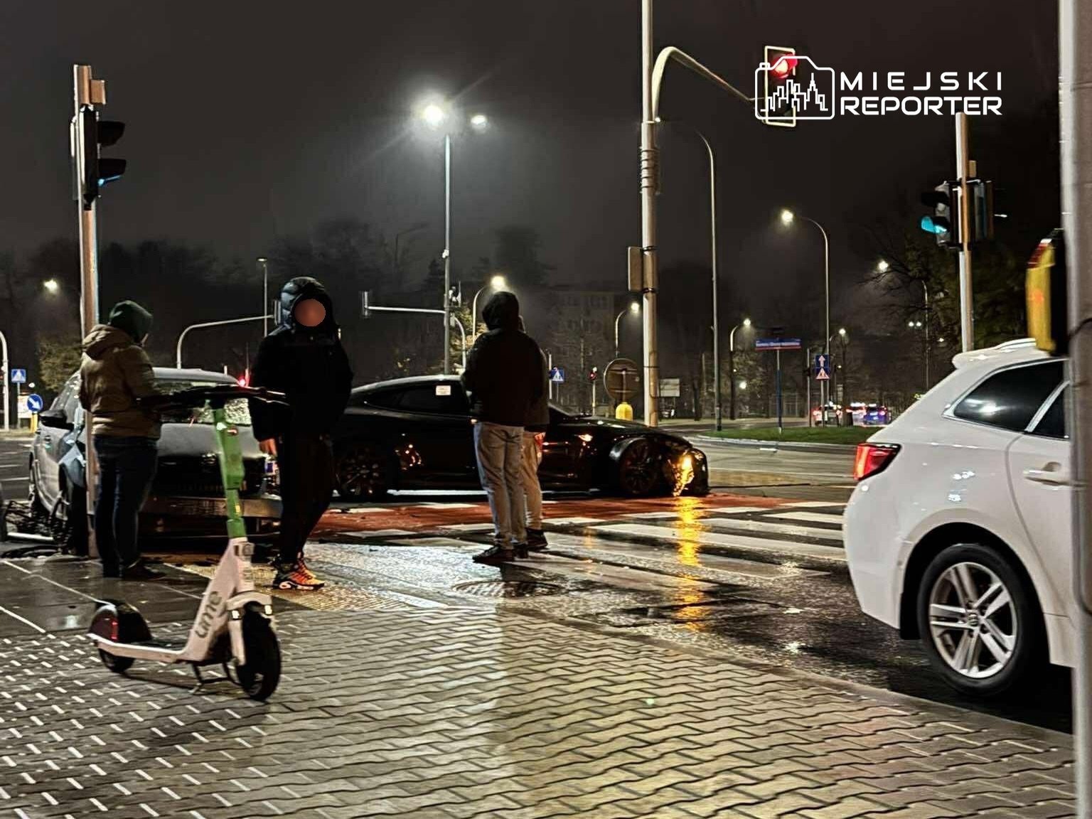 Grupa osób stoi przy skrzyżowaniu, obok zaparkowanego samochodu, w deszczową noc. Na chodniku widoczna hulajnoga elektryczna.