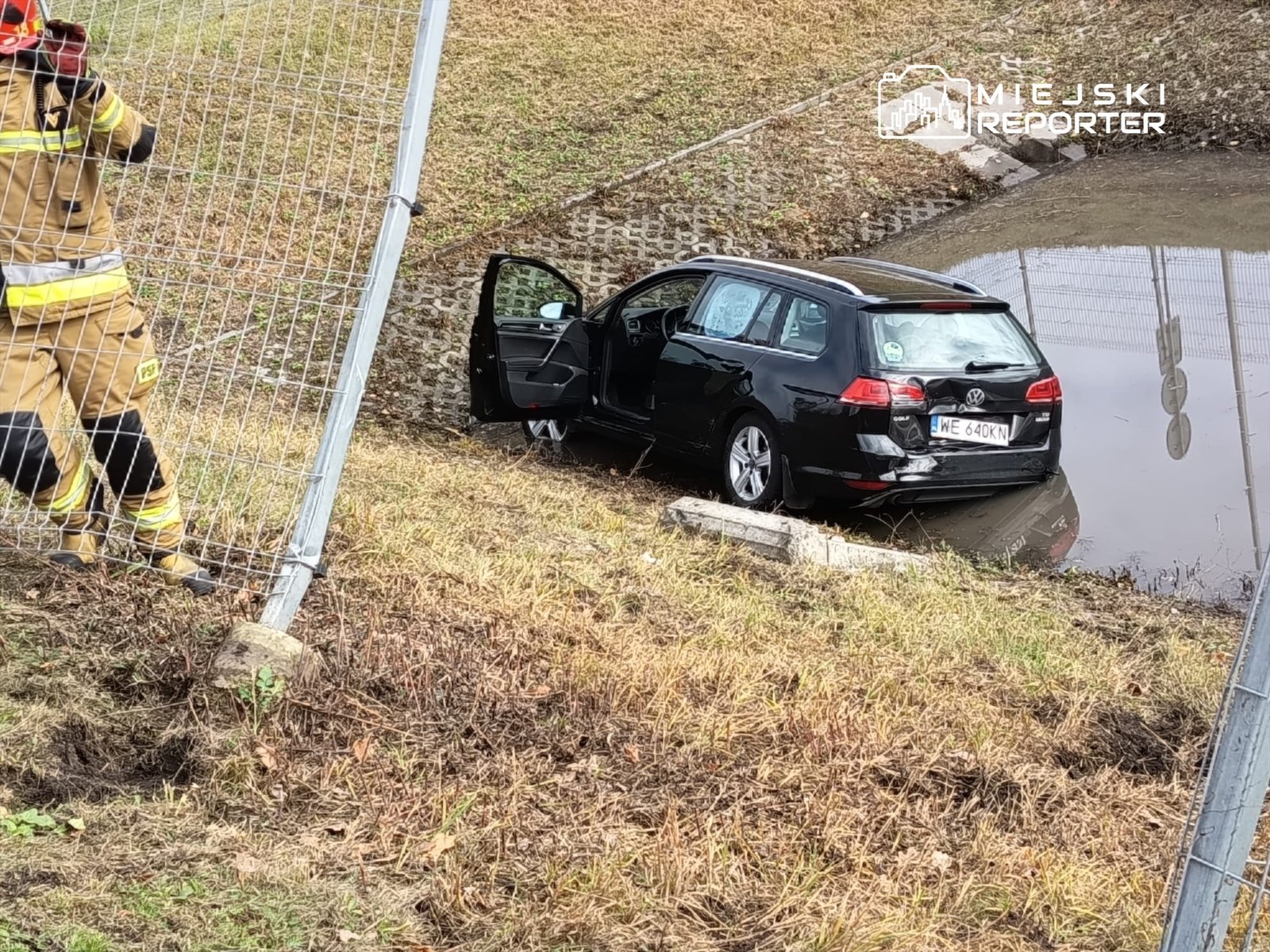 Strażak w mundurze obserwuje czarny samochód osobowy częściowo zanurzony w wodzie obok ogrodzenia.