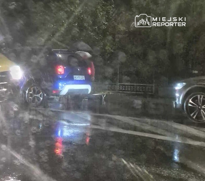 Niebieski samochód osobowy stoi na mokrej drodze, obok niego przejeżdża żółty pojazd. Deszczowa pogoda utrudnia widoczność.