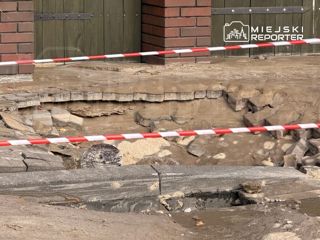 Uszkodzona nawierzchnia wokół wjazdu, odsłaniająca głęboką wyrwę, zabezpieczona taśmą ostrzegawczą.