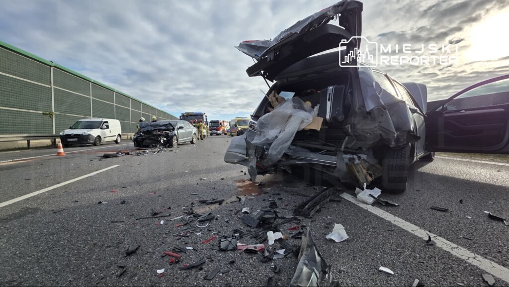 Zniszczony samochód na drodze, wokół leżą odłamki i fragmenty pojazdów, w tle strażacy udzielają pomocy.