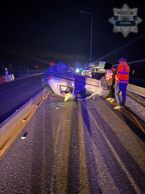 Samochód leży na dachu na jezdni, a obok niego stoją pracownicy w kamizelkach odblaskowych oraz policjanci.
