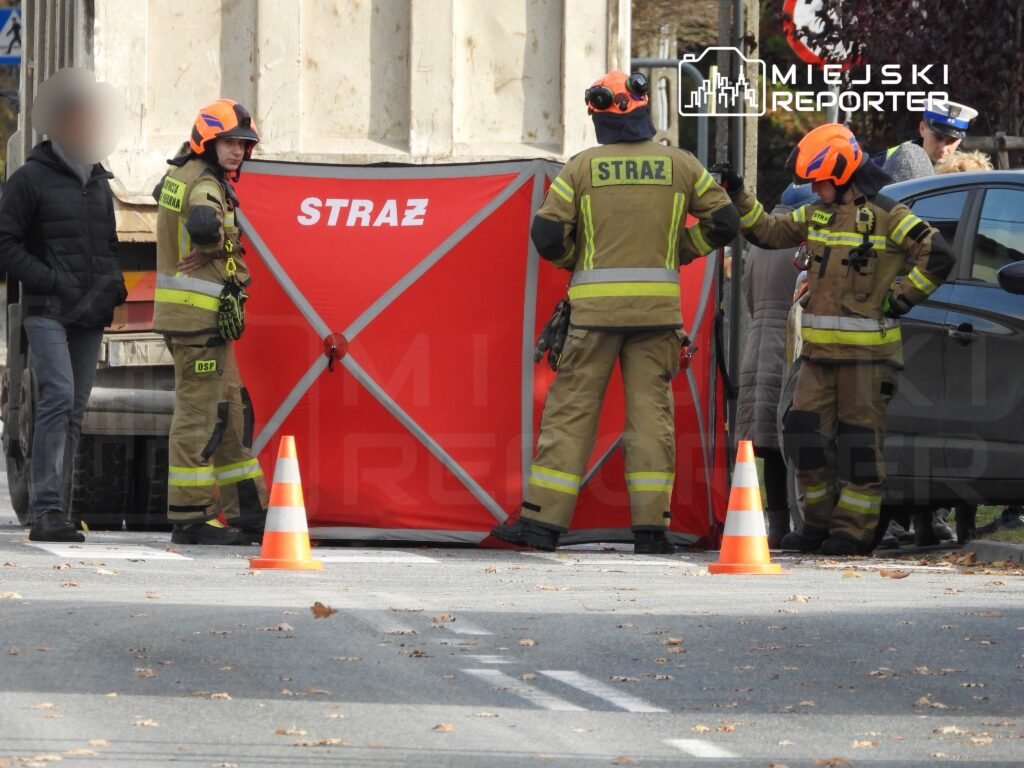 Czterej strażacy w hełmach rozmawiają przy czerwonym namiocie z napisem "STRAŻ" na poboczu drogi.