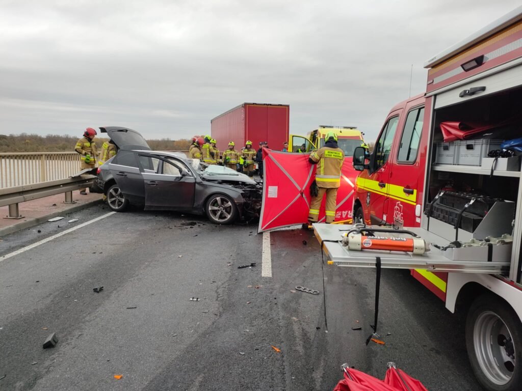 Czterech strażaków pracuje przy wypadku drogowym, obok uszkodzonego samochodu, na ruchliwej drodze.