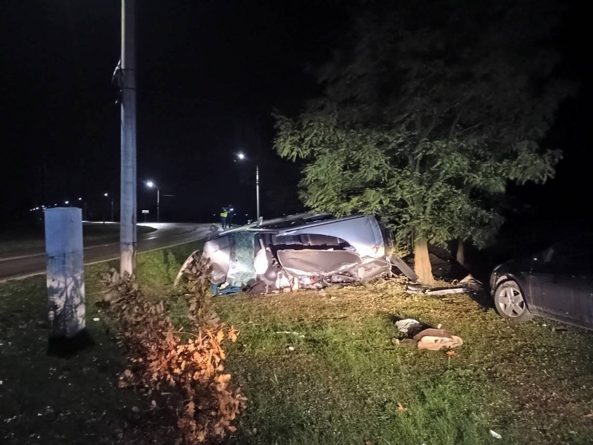Rozbity samochód osobowy leży na boku przy drodze, obok rosnącego drzewa, w nocy. W tle widoczne są latarnie uliczne.