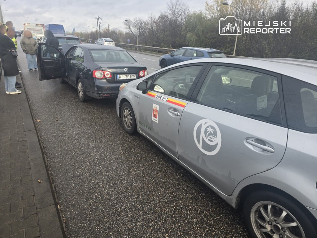 Trzy osoby stoją obok dwóch uszkodzonych samochodów na poboczu drogi, jeden z pojazdów ma otwarte drzwi.