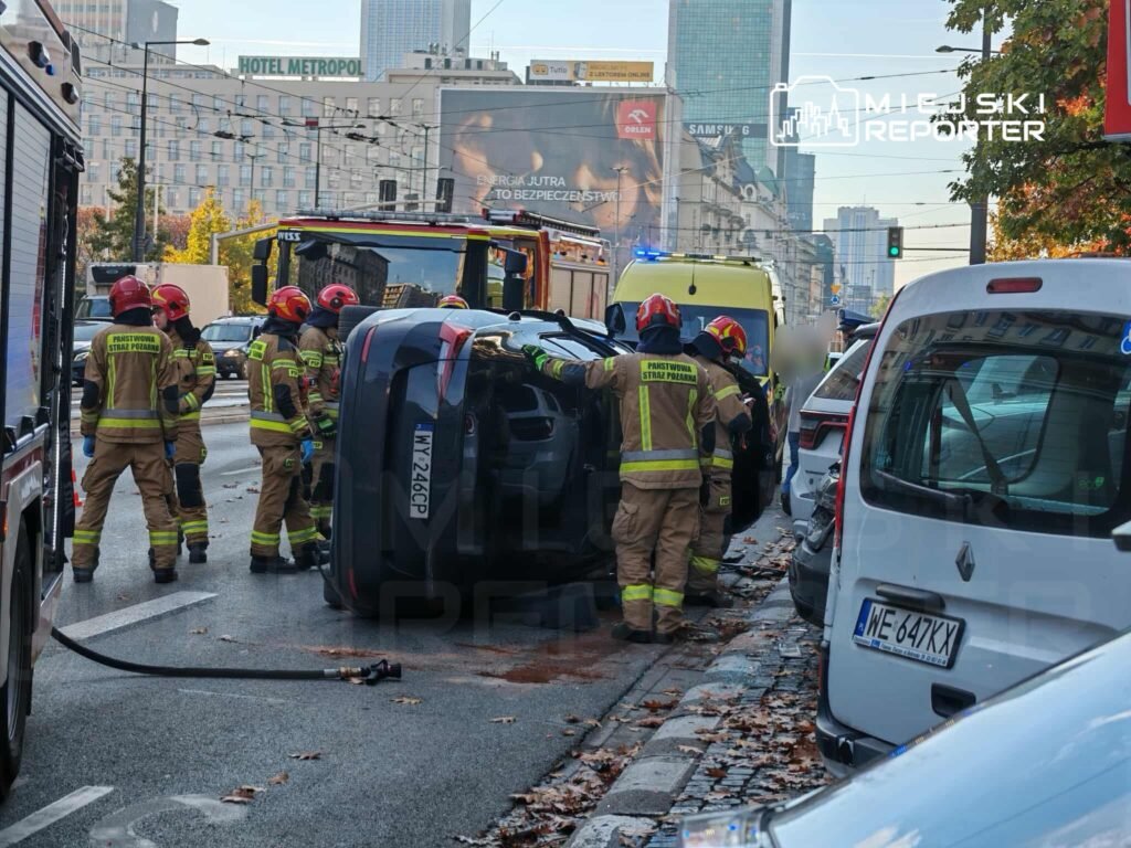 Grupa strażaków w mundurach pracuje przy przewróconym samochodzie na ruchliwej ulicy w mieście.