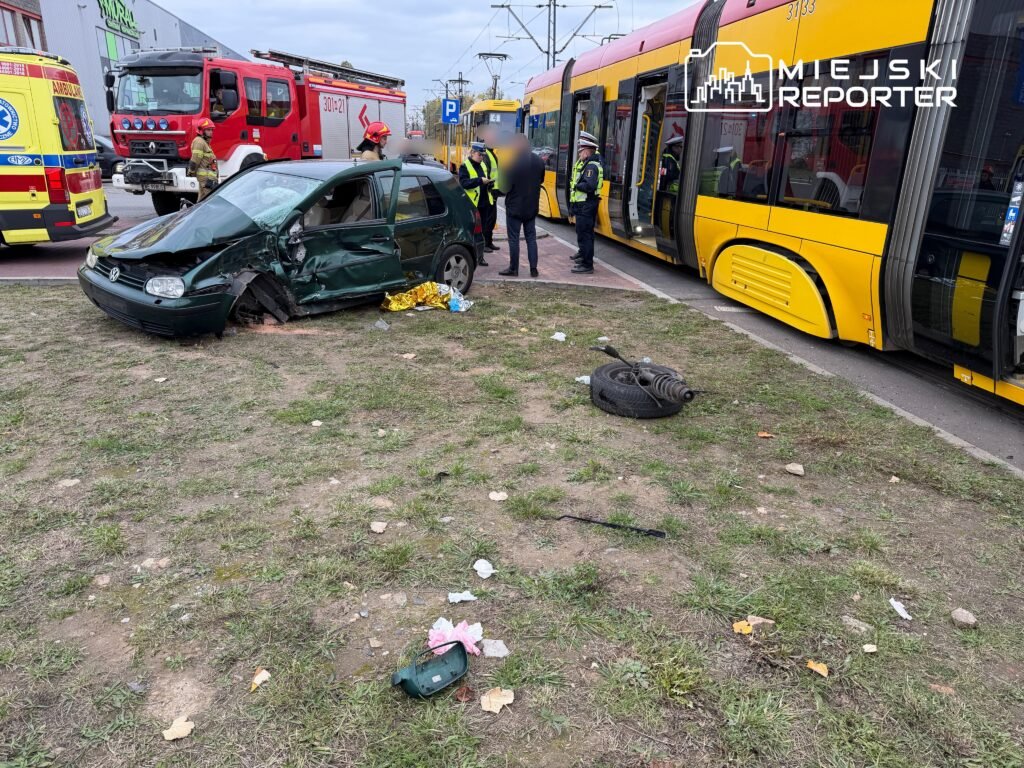 Zniszczony samochód osobowy stoi na trawie obok żółtego tramwaju, a strażacy i policjanci pomagają poszkodowanym.