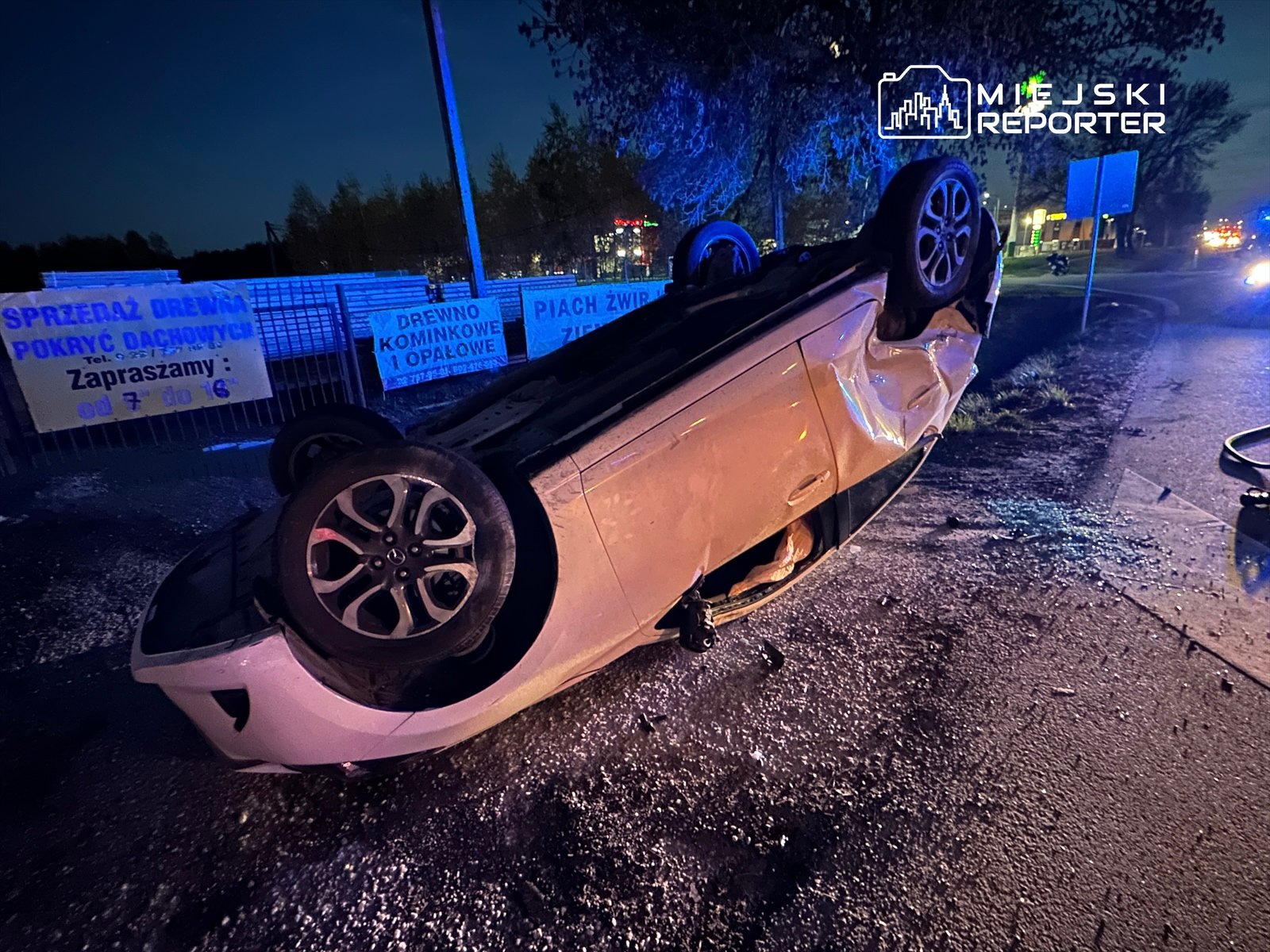 Przewrócony na dach samochód osobowy leży na jezdni, w tle widoczne reklamy i niebieskie światła policyjne.