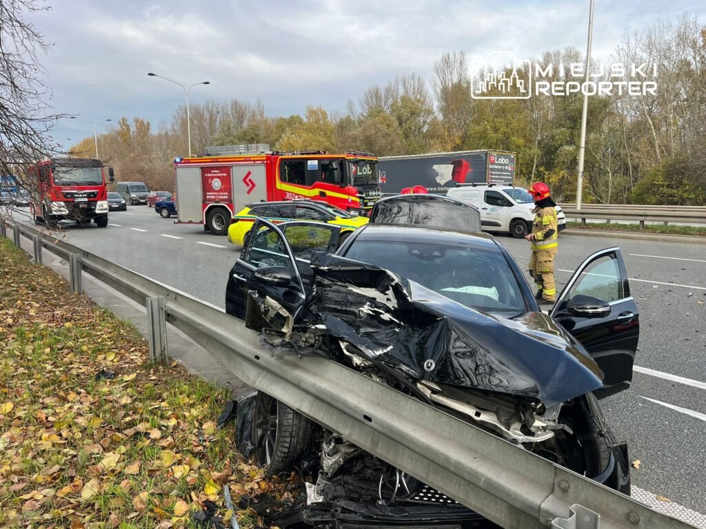 Czarny samochód osobowy z uszkodzoną tylną częścią stoi przy barierce drogi, obok interweniują strażacy i karetka.