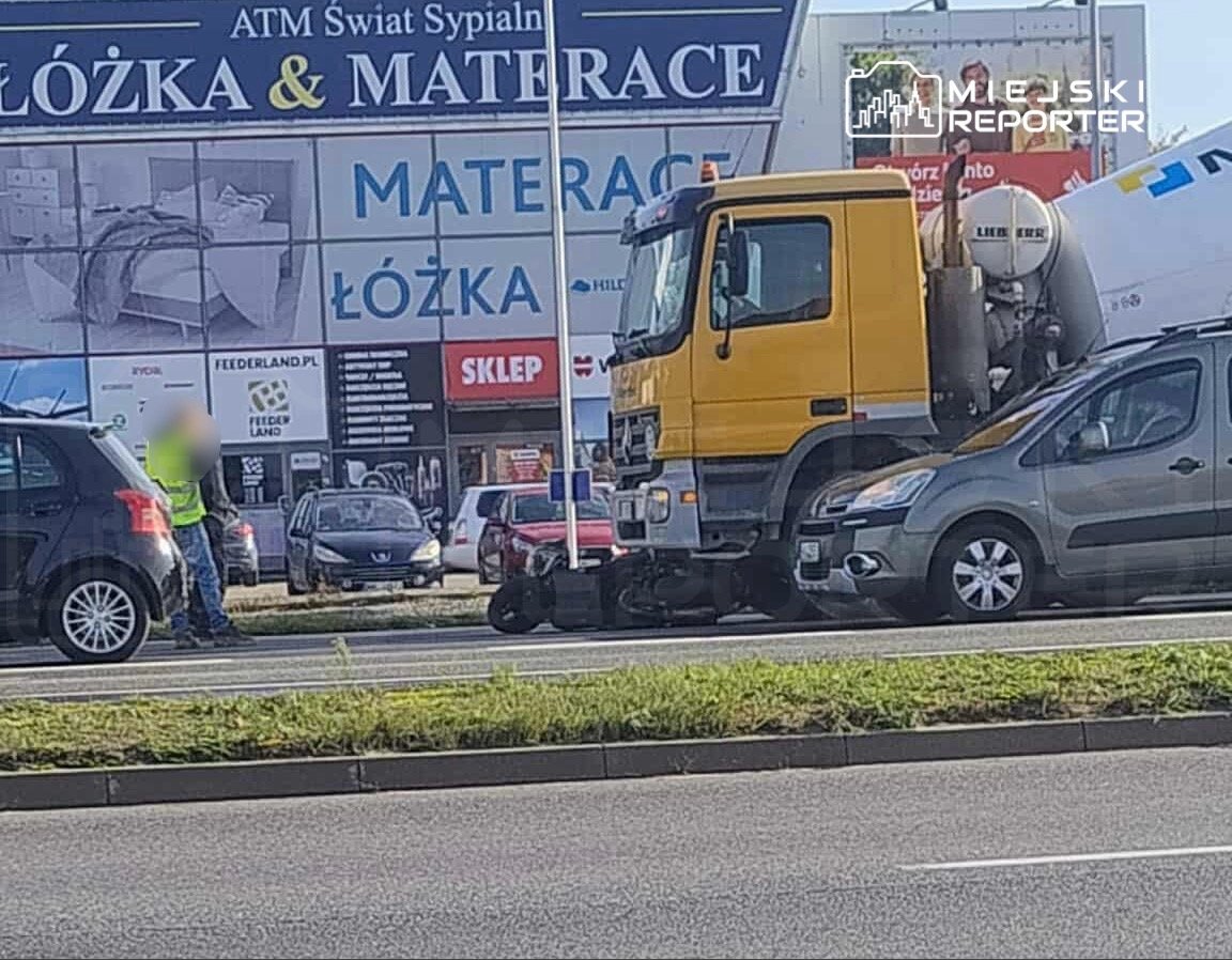 Mężczyzna w kamizelce odblaskowej rozmawia z kierowcą na ruchliwej ulicy obok dużego billboardu reklamowego.