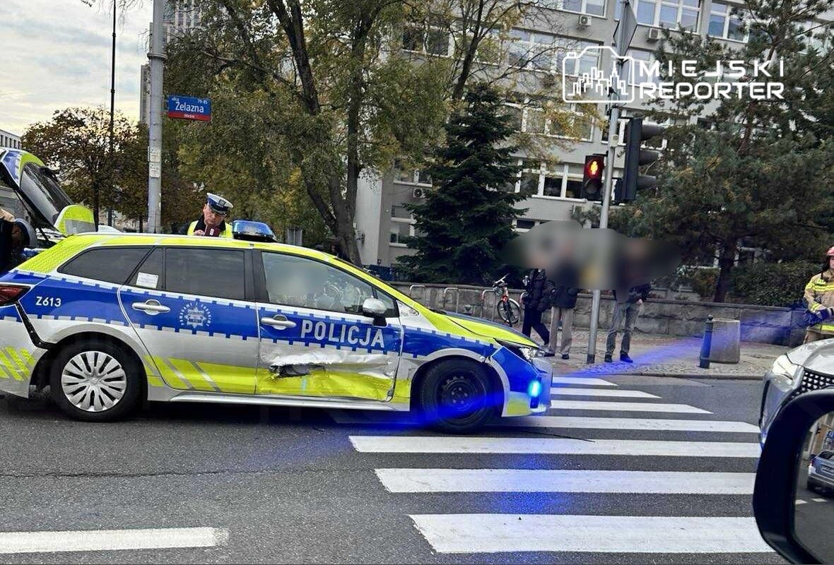 Uszkodzony radiowóz policyjny zatrzymany na przejściu dla pieszych, obok grupy osób i sygnalizacji świetlnej.
