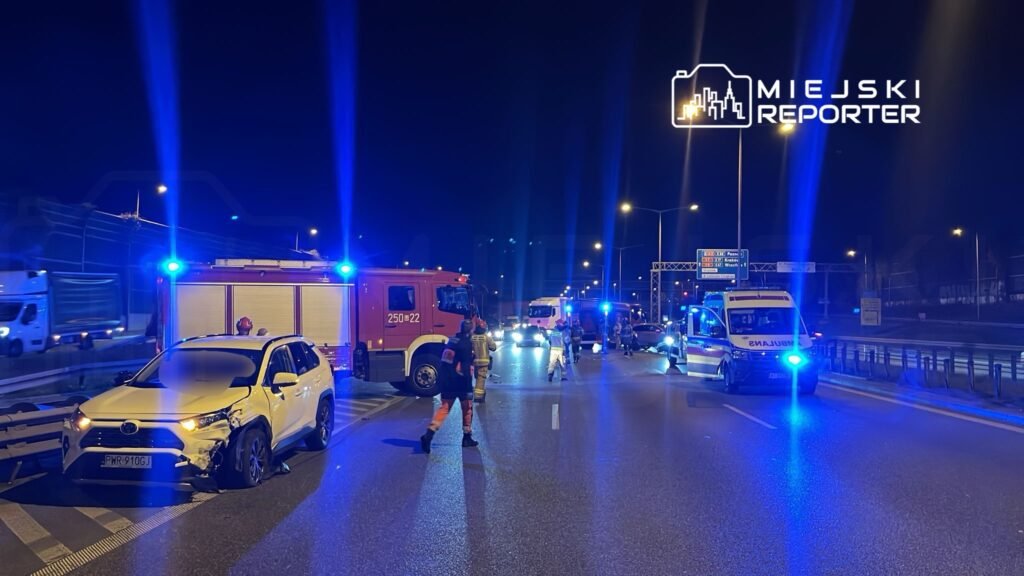 Na drodze nocą zderzone pojazdy, obok straż pożarna i karetka, w tle włączone sygnały świetlne.