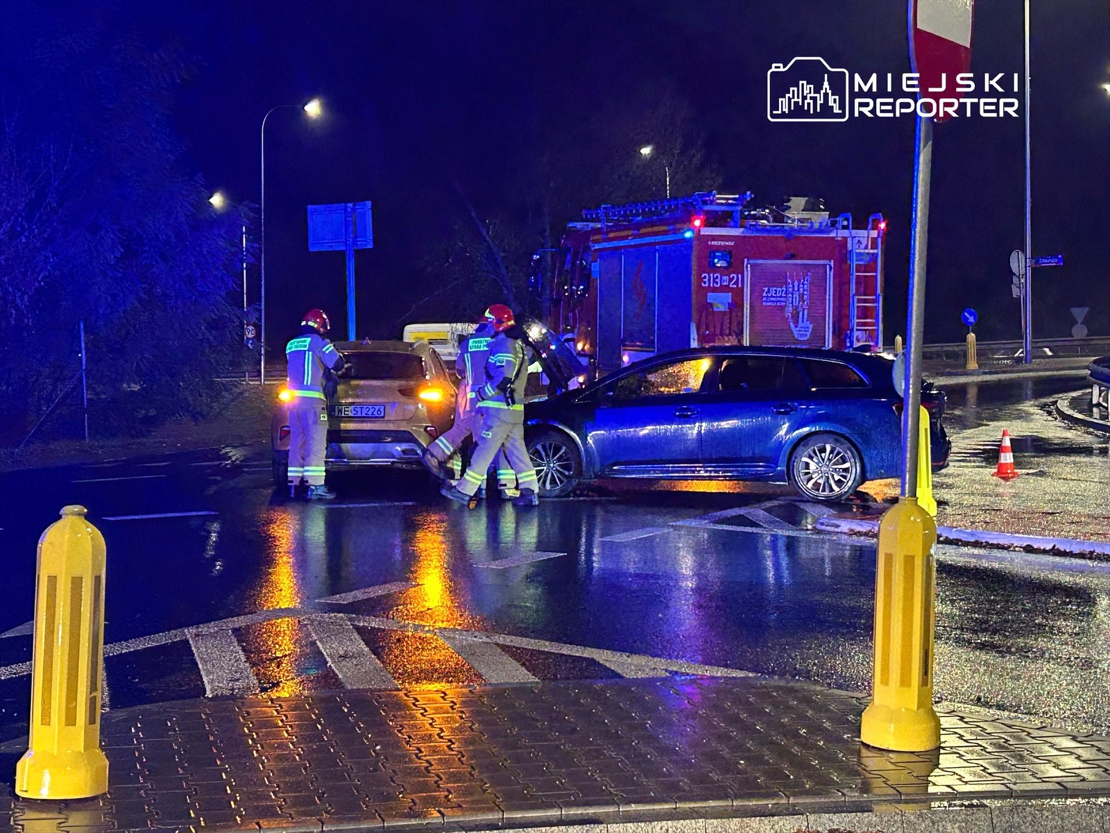 Trzech strażaków w kaskach bada uszkodzone samochody na mokrej jezdni w nocy, obok wozu strażackiego.