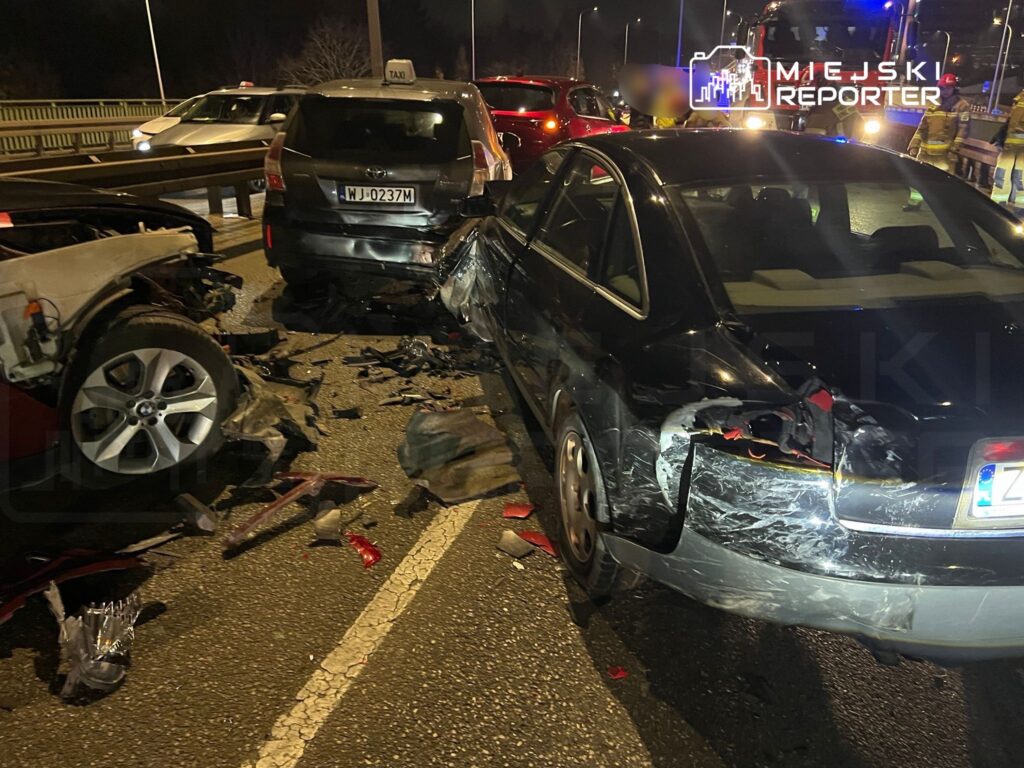 Zderzenie trzech samochodów na ruchliwej drodze, z widocznymi uszkodzeniami pojazdów i fragmentami wraku na jezdni.