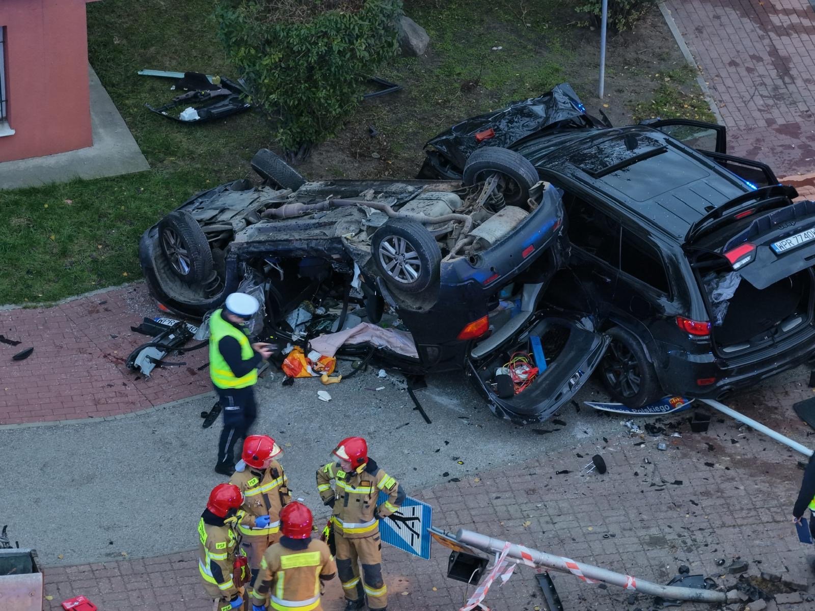 Grupa strażaków w hełmach ocenia skutki wypadku drogowego z przewróconym samochodem na chodniku.