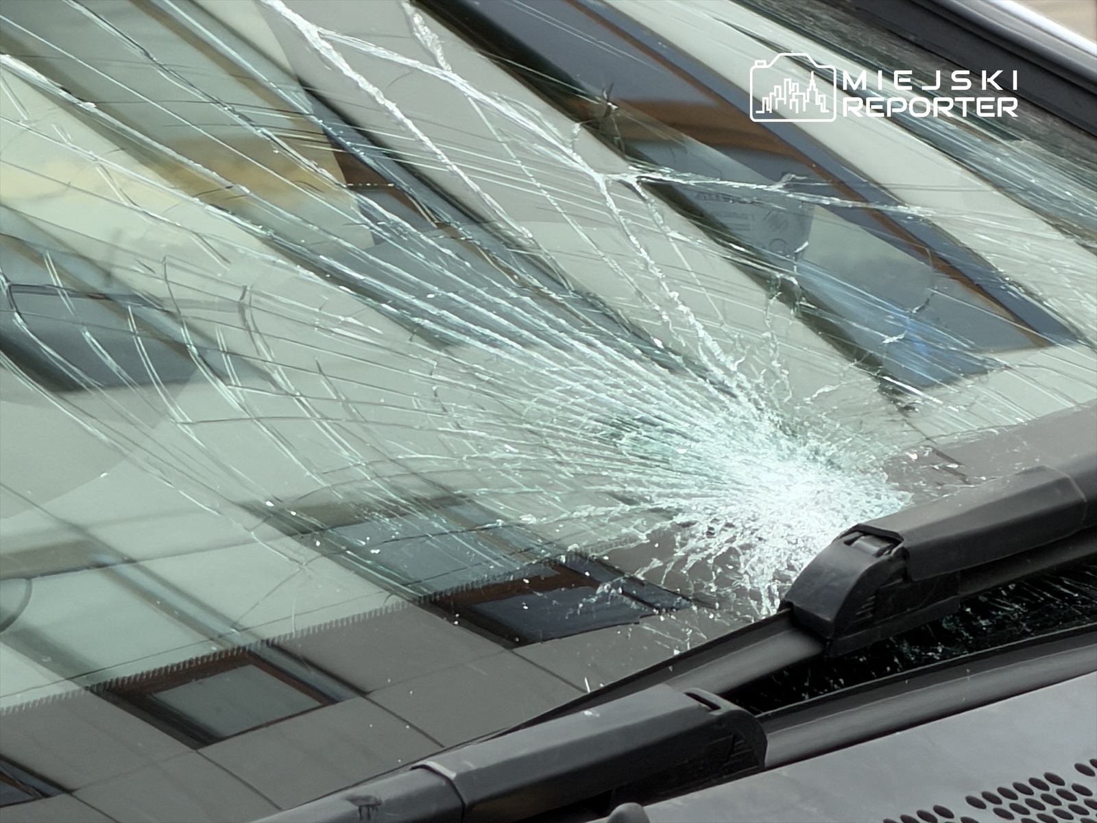 Szyba przednia samochodu z rozległym pęknięciem, w tle odbicia budynków.