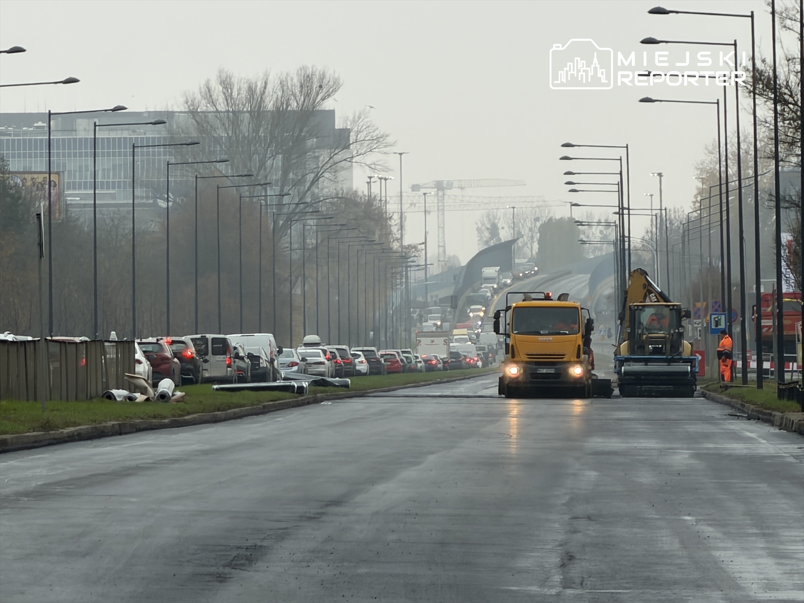 Ruch wstrzymany na ruchliwej ulicy, gdzie żółty samochód ciężarowy i maszyna budowlana pracują w otoczeniu korkujących się pojazdów.