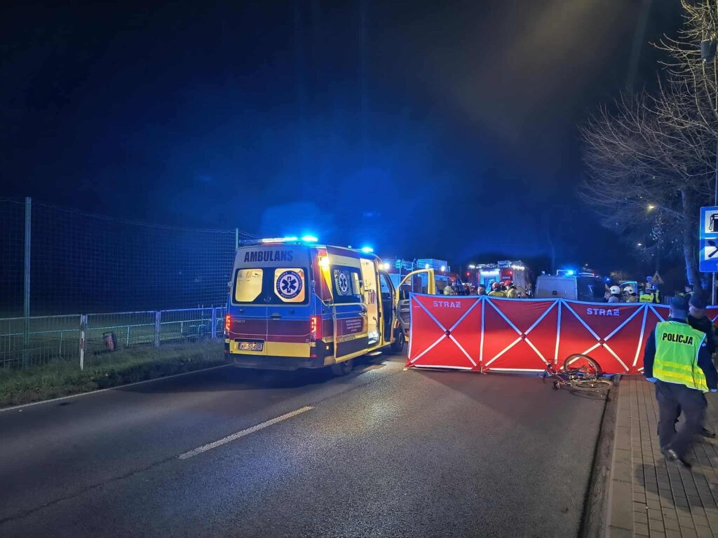 Ambulans z włączonymi sygnałami świetlnymi stoi na drodze, obok bariery zabezpieczającej miejsce zdarzenia, w nocy.