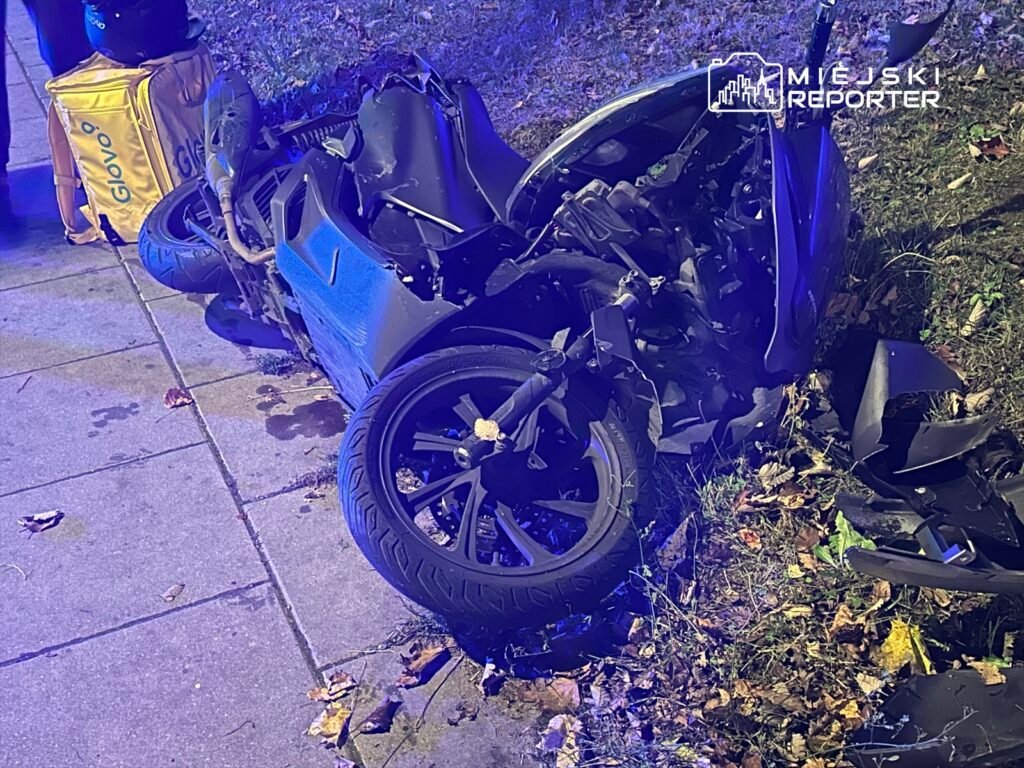 Zniszczony motocykl leży na chodniku, obok torba dostawcza. W tle widać zieleń i liście na ziemi.
