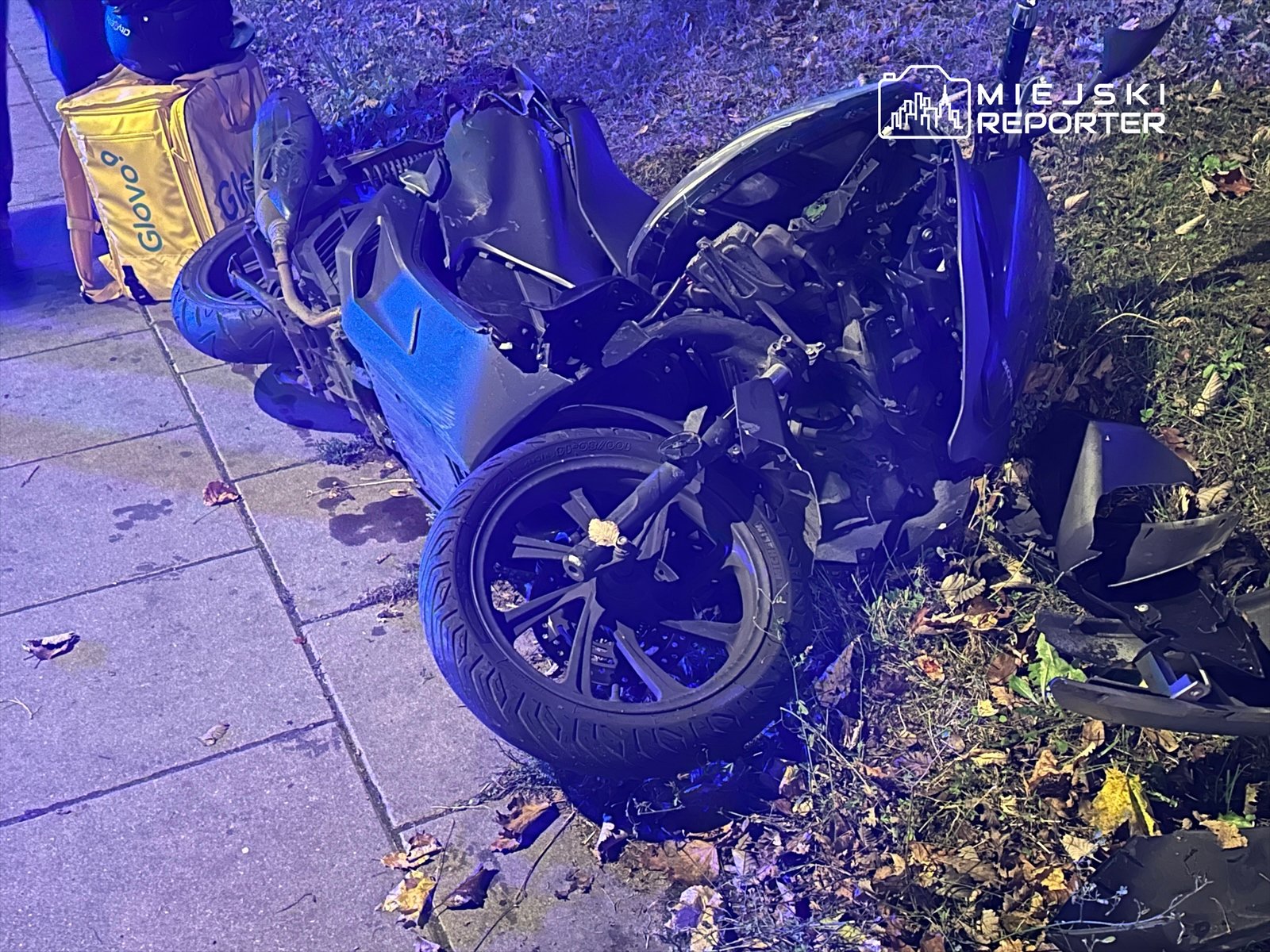 Zniszczony motocykl leży na chodniku, obok torba dostawcza. W tle widać zieleń i liście na ziemi.