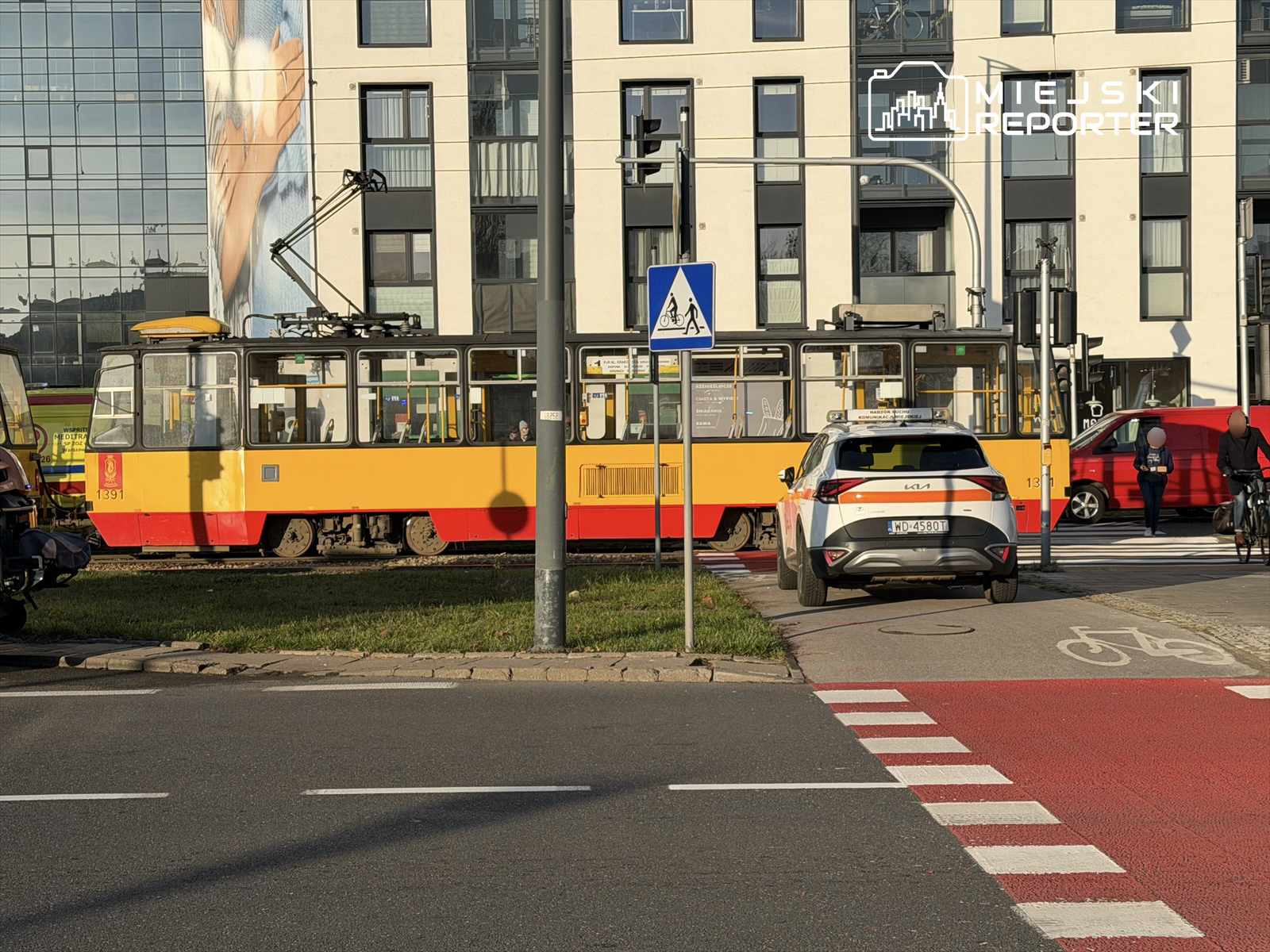 Tramwaj w żółto-czerwonych barwach zatrzymany na przystanku obok nowoczesnego budynku z dużymi oknami.
