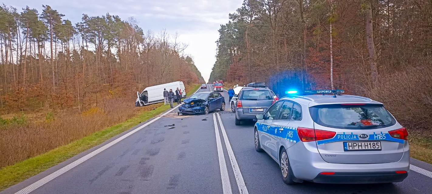 Wielu ludzi zbiera się przy miejscu wypadku drogowym, gdzie zderzyły się dwa pojazdy. Na drodze stoją radiowozy policyjne z włączonymi sygnałami świetlnymi.