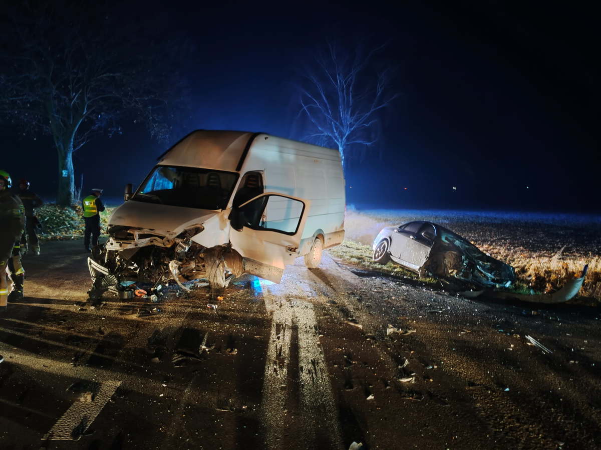 W nocy dwa rozbite pojazdy, dostawczy i osobowy, leżą na drodze, a obok pracują strażacy i policjanci.