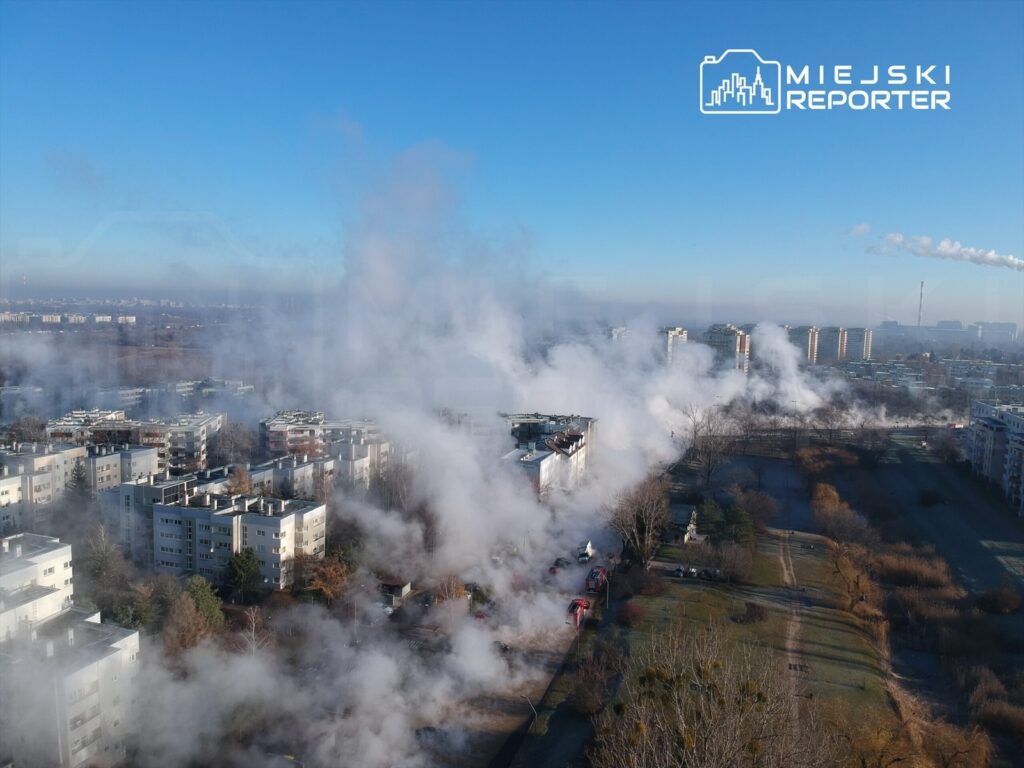 Widok na osiedle mieszkalne z unoszącym się dymem w tle, w słoneczny dzień z błękitnym niebem.