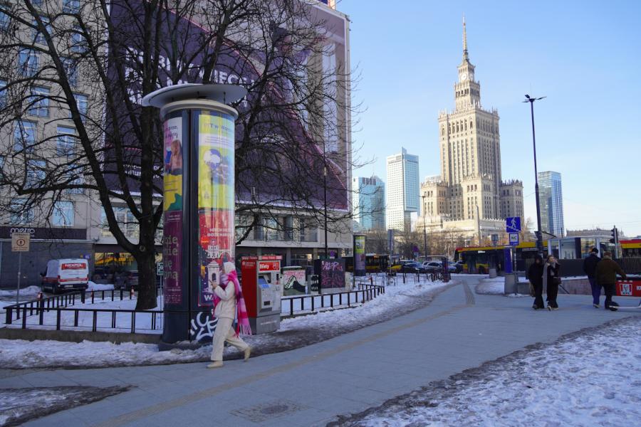 Mężczyzna w białym kombinezonie z różowym szalikiem spacerujący po ośnieżonej ulicy z widokiem na wieżowiec.