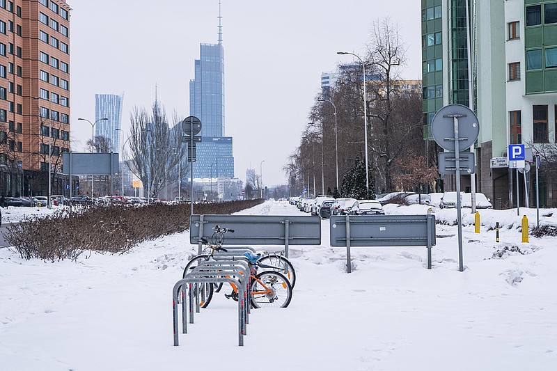 Zimowy krajobraz z rowerami przypiętymi do stojaka na zaśnieżonej ulicy, w tle nowoczesne wieżowce.