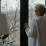 Starsza kobieta patrzy przez okno w swoim mieszkaniu