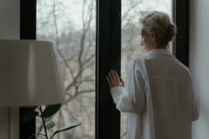 Starsza kobieta patrzy przez okno w swoim mieszkaniu